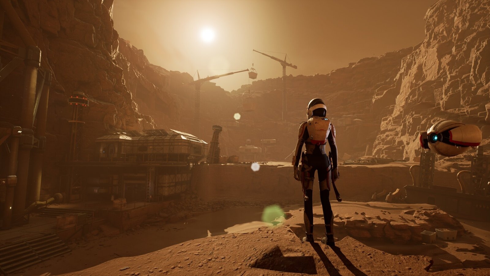 A suited astronaut and a hovering drone look out over a Mars colony under construction, cranes silhouetted against a low sun.